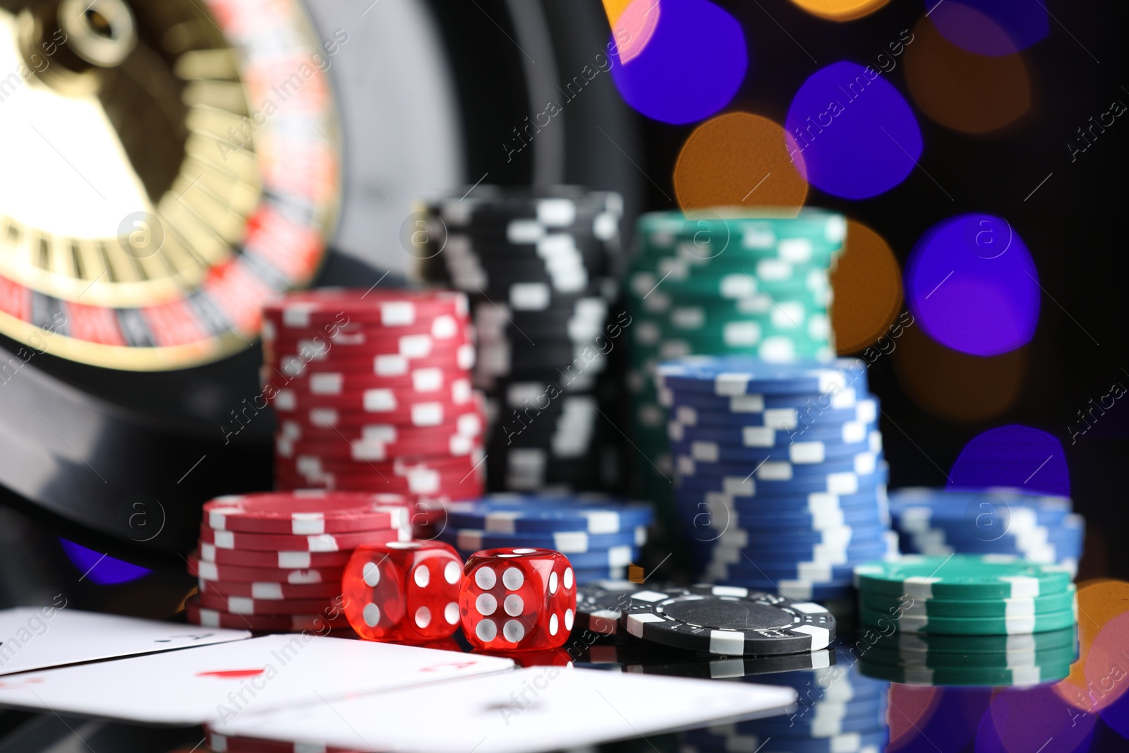 Photo of Casino chips, playing cards, roulette wheel and dice on mirror surface against blurred lights, closeup