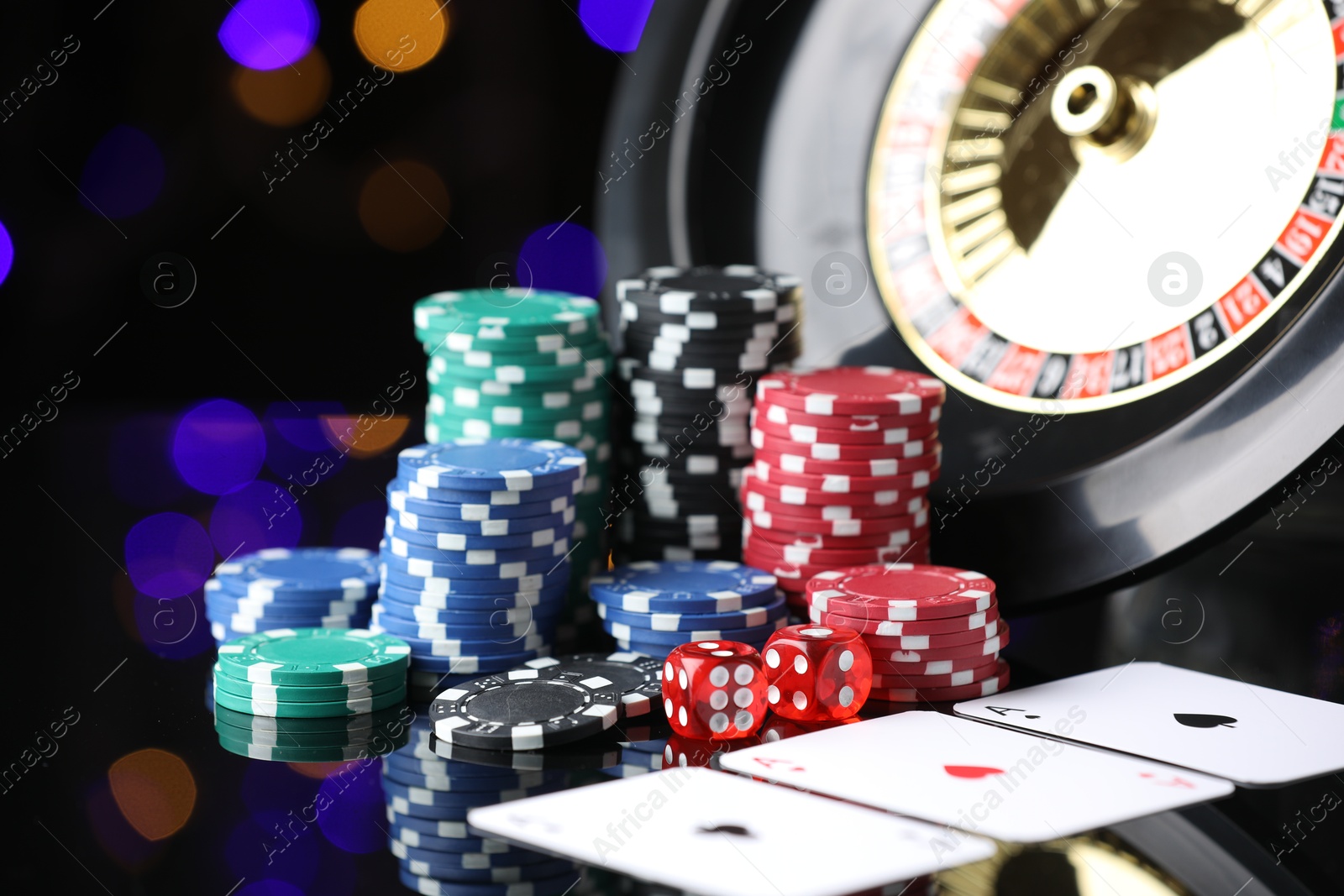 Casino chips, playing cards, roulette wheel and dice on mirror surface against blurred lights, closeup Photo of Casino chips, playing cards, roulette wheel and dice on mirror surface against blurred lights, closeup