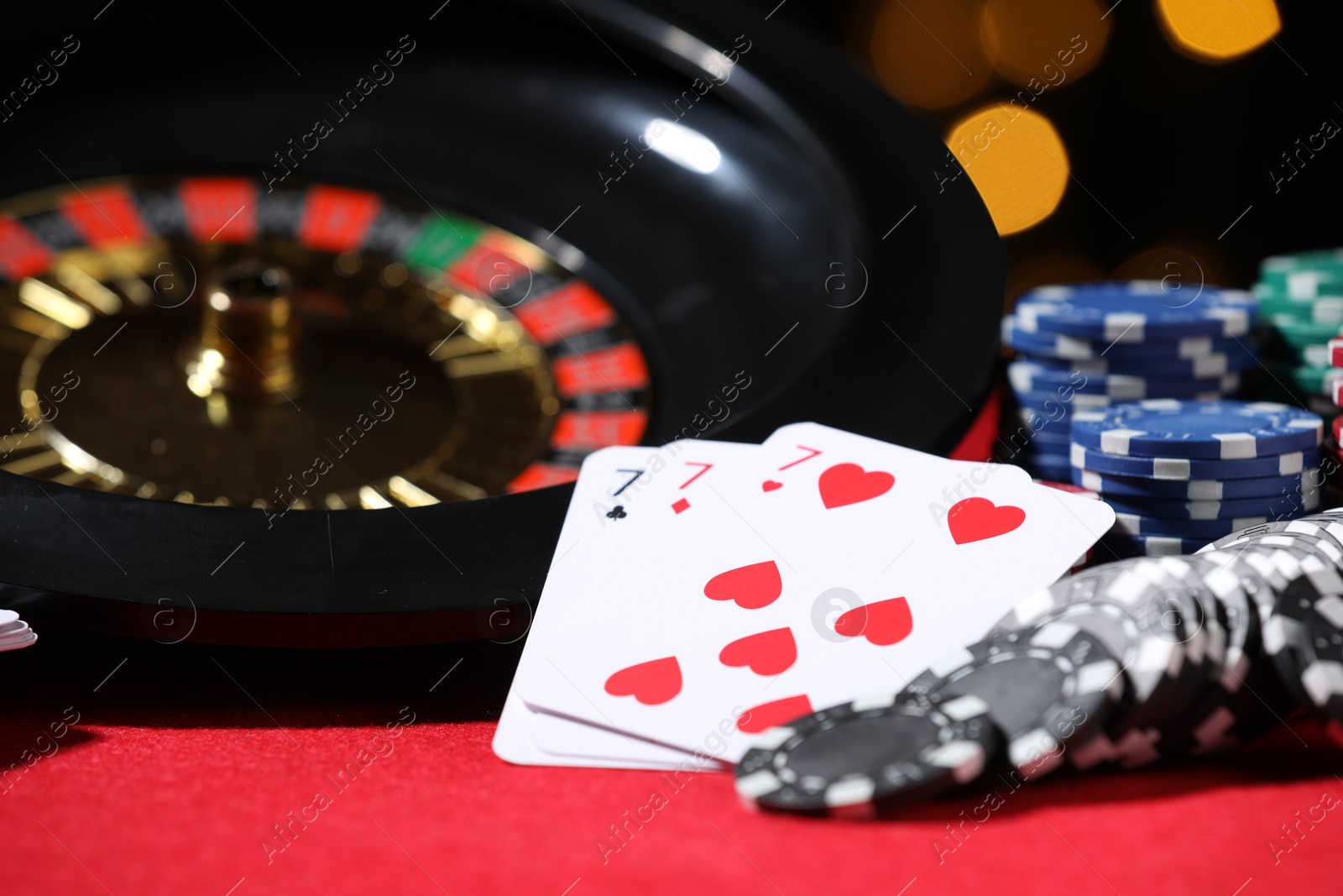 Casino chips, playing cards and roulette wheel on red table against blurred lights, closeup Photo of Casino chips, playing cards and roulette wheel on red table against blurred lights, closeup