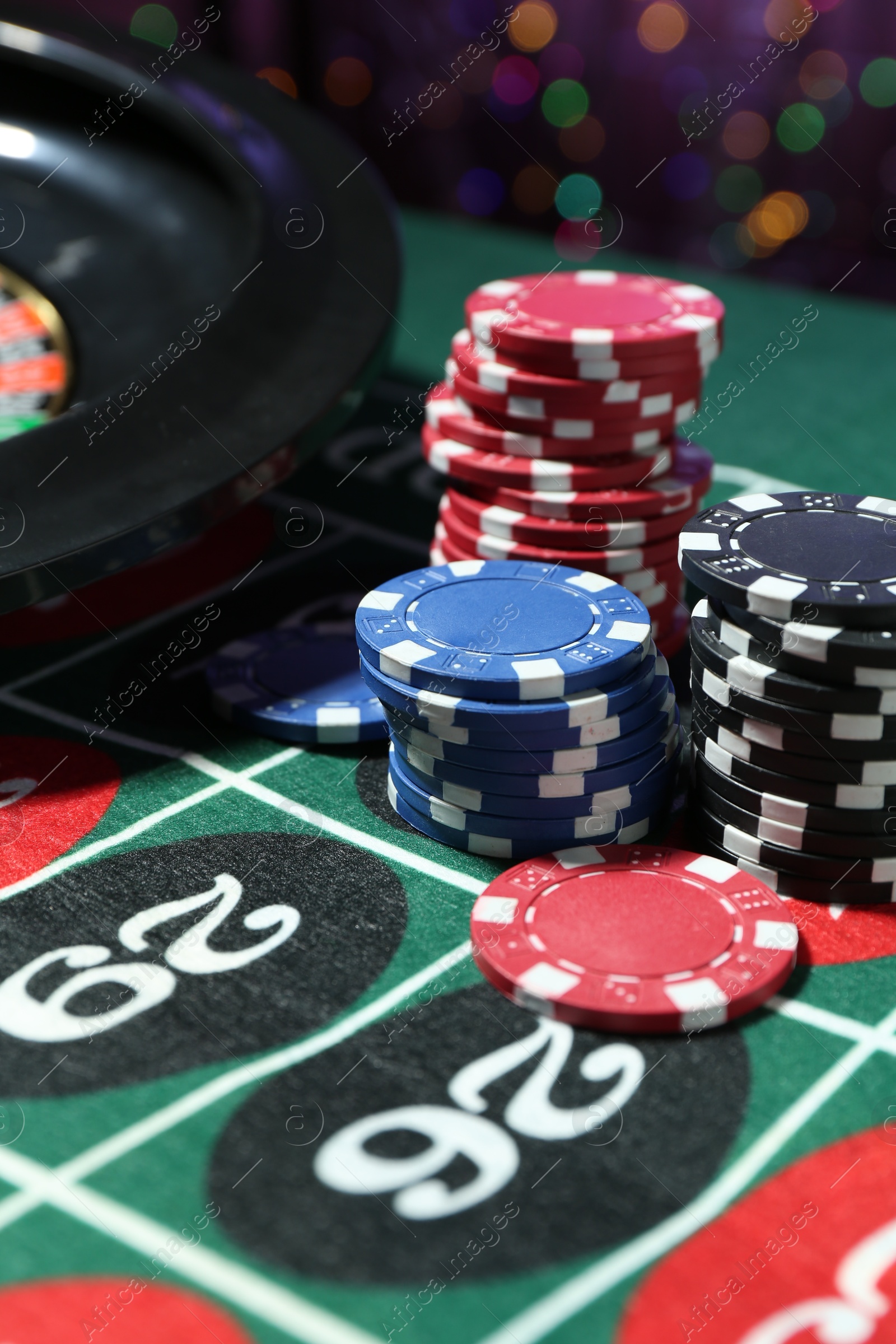 Photo of Casino chips and roulette wheel on table against blurred lights, closeup