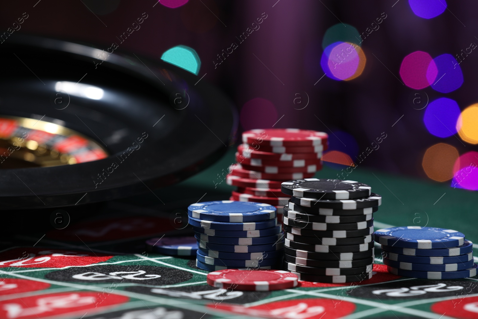 Casino chips and roulette wheel on table against blurred lights, closeup Photo of Casino chips and roulette wheel on table against blurred lights, closeup