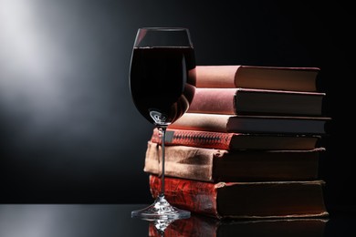 Photo of Tasty wine in glass and stack of books on mirror table against dark background, closeup. Space for text