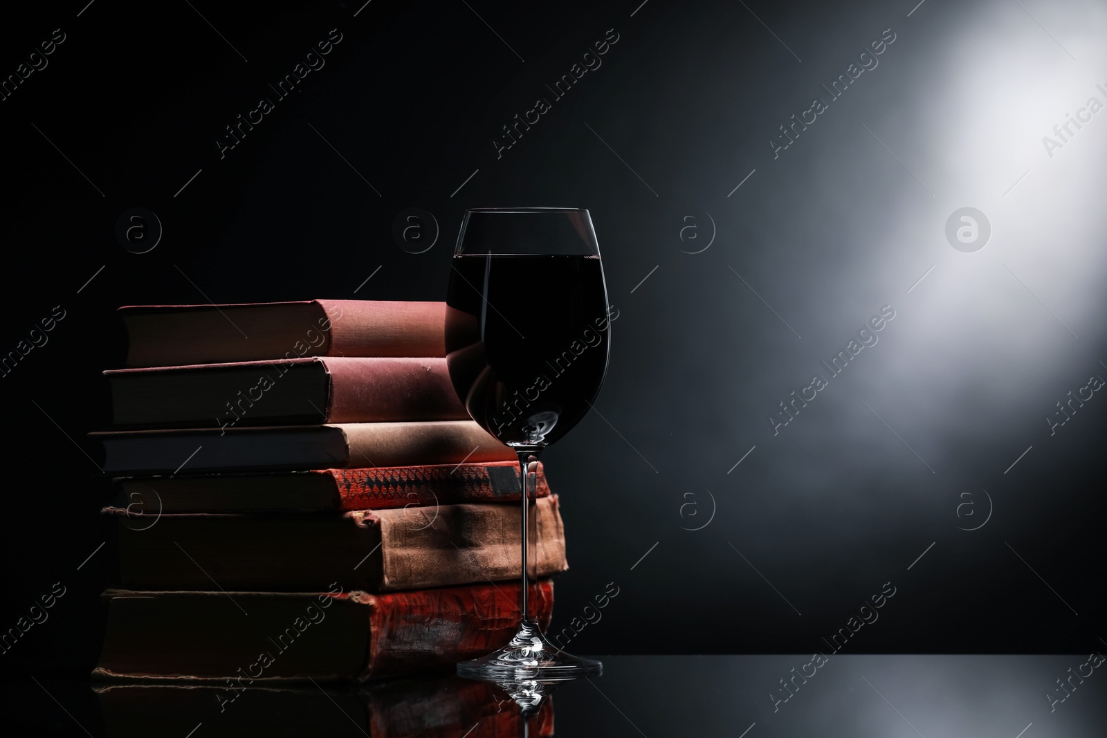 Tasty wine in glass and stack of books on mirror table against dark background. Space for text Photo of Tasty wine in glass and stack of books on mirror table against dark background. Space for text