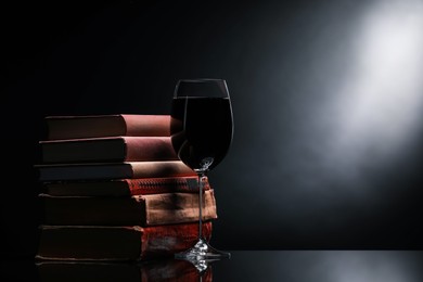 Photo of Tasty wine in glass and stack of books on mirror table against dark background. Space for text