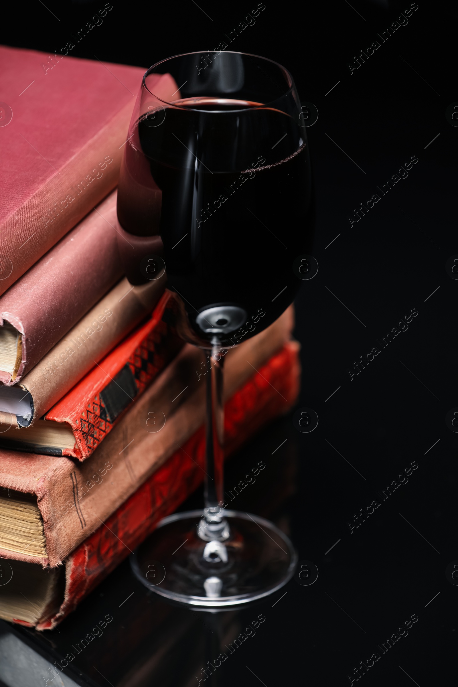 Red wine in glass and stack of books on mirror table against black background, closeup Photo of Red wine in glass and stack of books on mirror table against black background, closeup