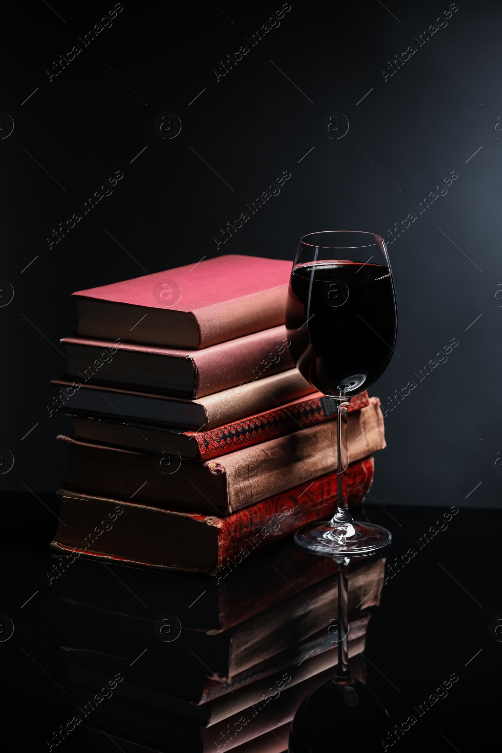 Red wine in glass and stack of books on mirror table against black background Photo of Red wine in glass and stack of books on mirror table against black background
