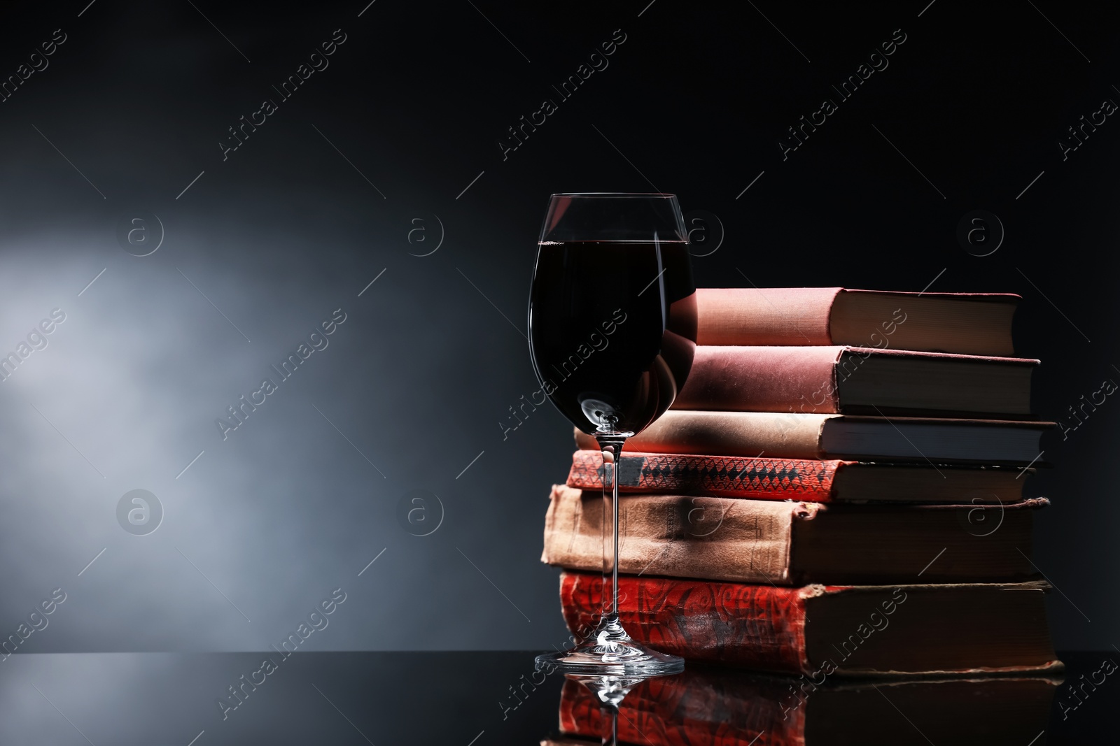 Tasty wine in glass and stack of books on mirror table against dark background. Space for text Photo of Tasty wine in glass and stack of books on mirror table against dark background. Space for text
