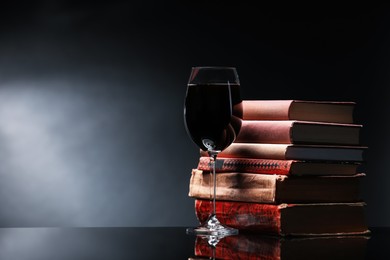 Photo of Tasty wine in glass and stack of books on mirror table against dark background. Space for text