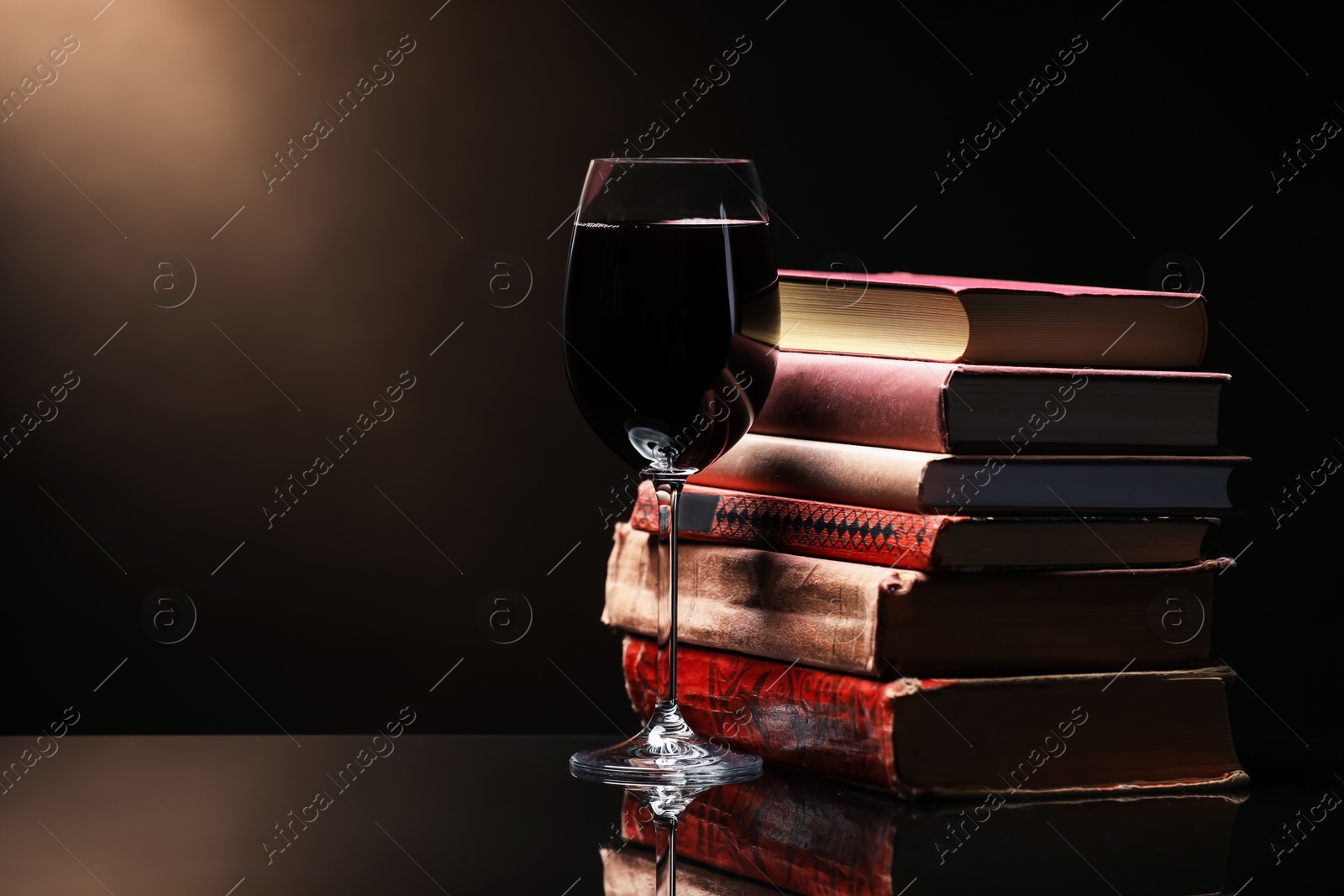 Photo of Tasty wine in glass and stack of books on mirror table against dark background. Space for text