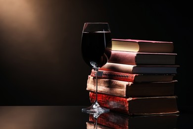 Photo of Tasty wine in glass and stack of books on mirror table against dark background. Space for text