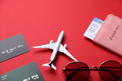 Photo of Credit cards, passport, flight ticket, airplane model and sunglasses on red background, above view