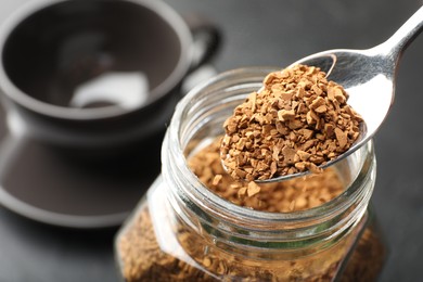 Photo of Adding granulated instant coffee from spoon into jar on dark table, closeup