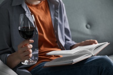 Photo of Woman with glass of wine reading book on sofa at home, closeup