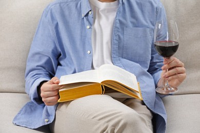 Photo of Woman with glass of wine reading book on sofa at home, closeup