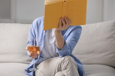 Photo of Woman with glass of wine reading book on sofa at home, closeup