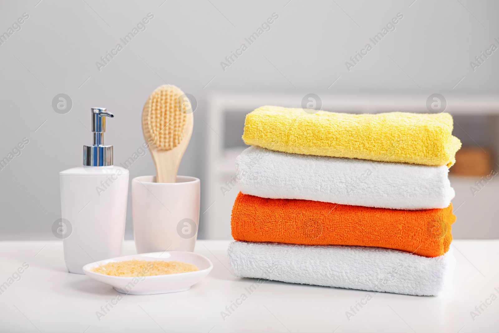 Stack of colorful towels, sea salt, liquid soap and brush on white table indoors Photo of Stack of colorful towels, sea salt, liquid soap and brush on white table indoors