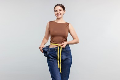 Photo of Beautiful woman with measuring tape wearing old jeans after weight loss on light grey background