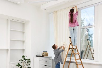 Photo of Couple putting up curtains using ladder in room