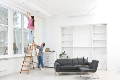 Photo of Couple putting up curtains using ladder in room