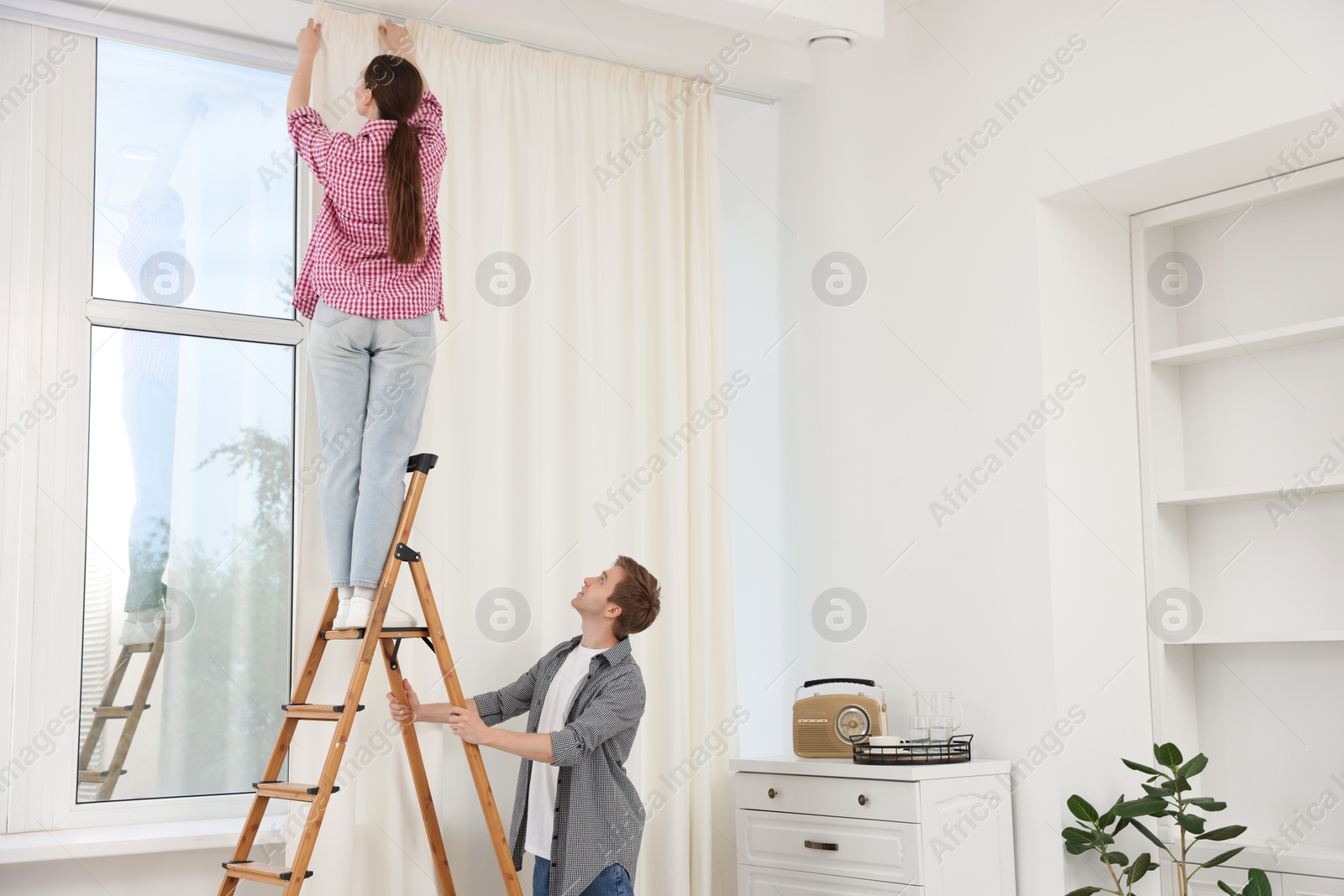 Couple putting up curtains using ladder in room Photo of Couple putting up curtains using ladder in room