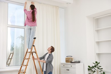 Photo of Couple putting up curtains using ladder in room
