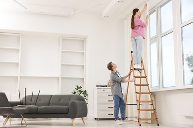 Photo of Couple putting up curtains using ladder in room