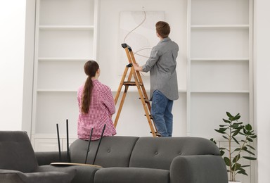 Photo of Couple putting painting on wall using ladder at home