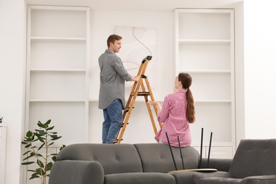 Photo of Couple putting painting on wall using ladder at home