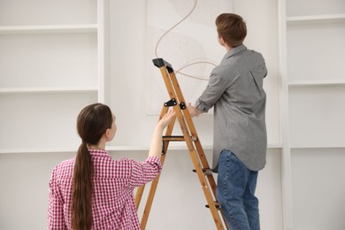 Photo of Couple putting painting on wall using ladder at home