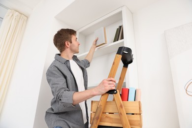 Photo of Man putting books on shelves using ladder at home, low angle view