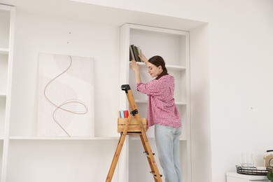 Photo of Woman putting books on shelves using ladder at home