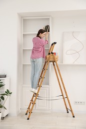 Photo of Woman putting books on shelves using ladder at home