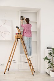 Photo of Woman putting books on shelves using ladder at home
