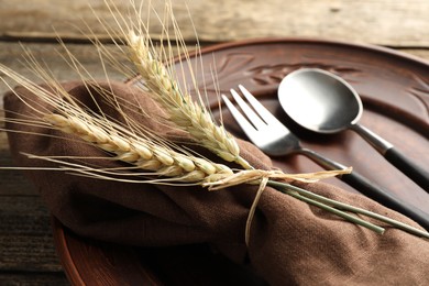 Photo of Wheat spikes, plate and cutlery on wooden table, closeup