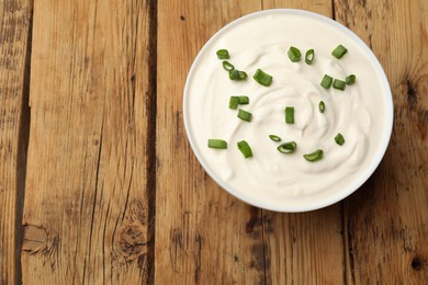 Photo of Tasty sour cream with green onions in bowl on wooden table, top view. Space for text