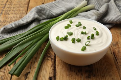 Photo of Tasty sour cream in bowl and green onions on wooden table, closeup