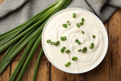Photo of Tasty sour cream in bowl and green onions on wooden table, flat lay