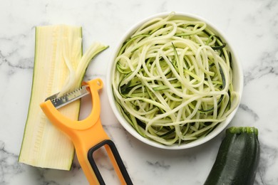 Photo of Raw zucchini noodles, peeler and fresh vegetables on white marble table, flat lay. Vegetarian dish