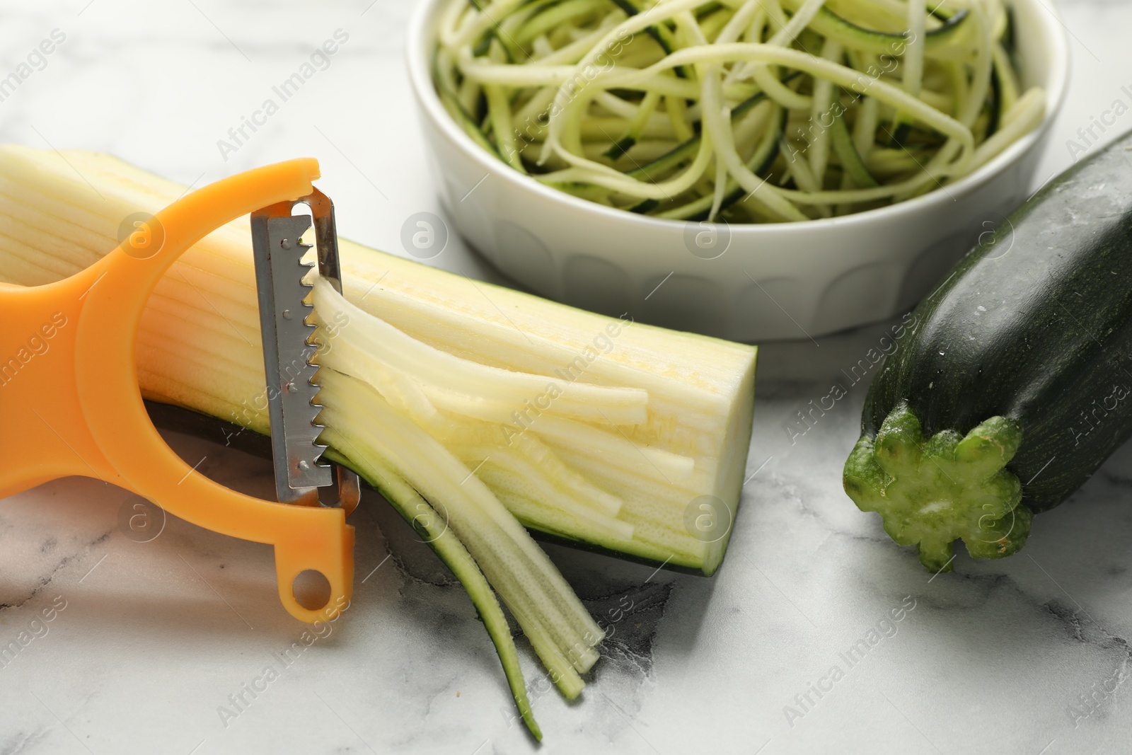 Photo of Raw zucchini noodles, peeler and fresh vegetables on white marble table, closeup. Vegetarian dish