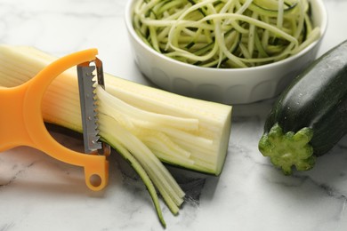 Photo of Raw zucchini noodles, peeler and fresh vegetables on white marble table, closeup. Vegetarian dish