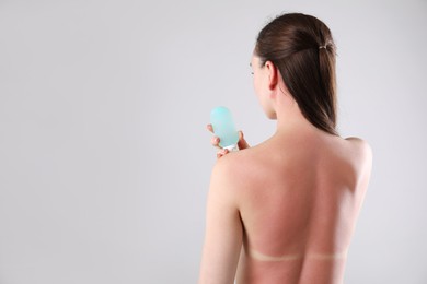 Photo of Woman with sunburned skin holding bottle of sun protection product on grey background, back view. Space for text
