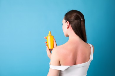 Photo of Woman with sunburned skin holding bottle of sun protection product on light blue background, back view. Space for text