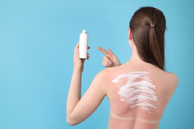 Photo of Woman with sunburned skin holding bottle of sun protection product on light blue background, back view. Space for text