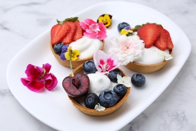 Photo of Sweet tartlets with berries and flowers on light marble table, closeup. Delicious dessert
