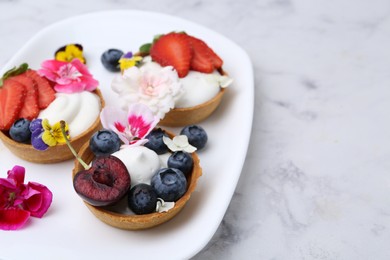 Photo of Sweet tartlets with berries and flowers on light marble table, closeup. Delicious dessert