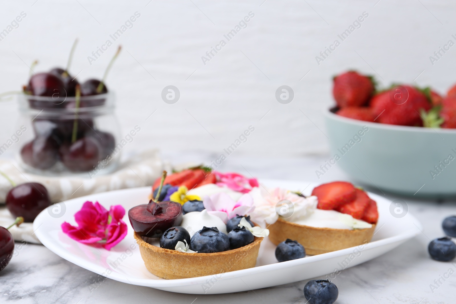 Photo of Sweet tartlets with berries and flowers on light marble table against white background, closeup. Delicious dessert