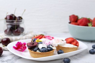 Photo of Sweet tartlets with berries and flowers on light marble table against white background, closeup. Delicious dessert