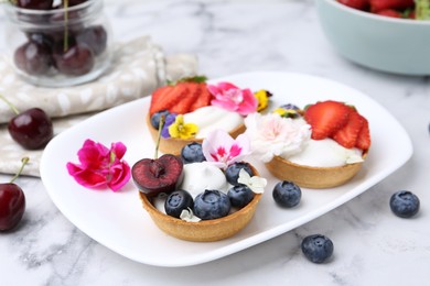 Photo of Sweet tartlets with berries and flowers on light marble table, closeup. Delicious dessert
