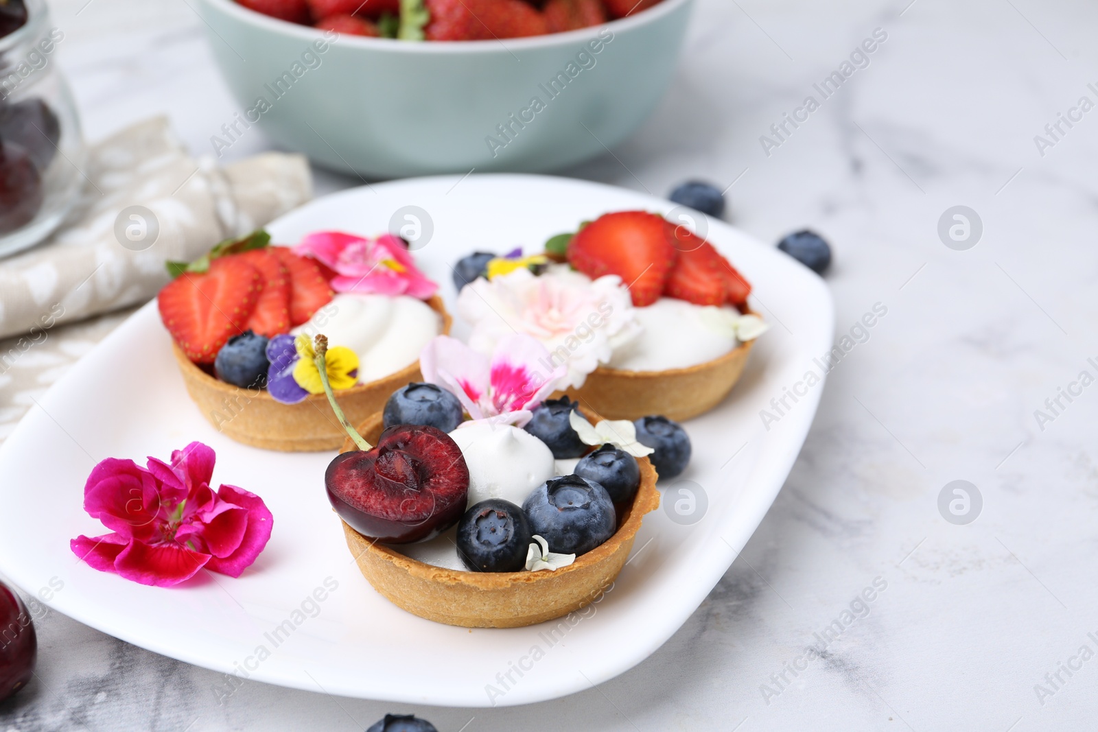 Sweet tartlets with berries and flowers on light marble table, closeup. Delicious dessert Photo of Sweet tartlets with berries and flowers on light marble table, closeup. Delicious dessert