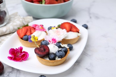 Photo of Sweet tartlets with berries and flowers on light marble table, closeup. Delicious dessert